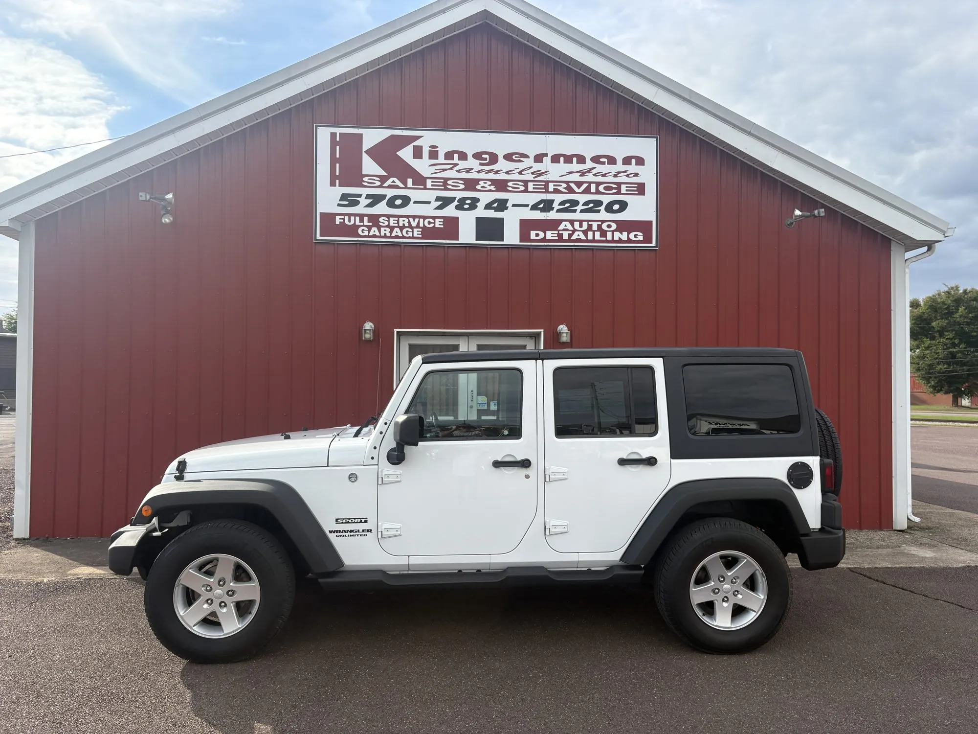 A white 2014 Jeep Wrangler Unlimited Sport with a black hardtop, parked in front of the Klingerman Family Auto building